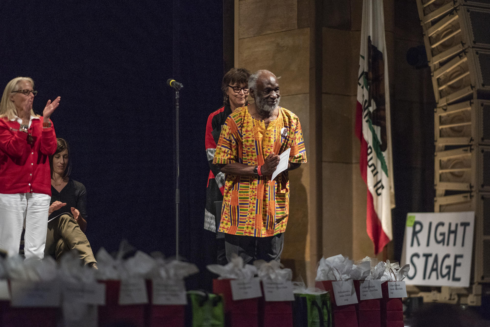 President Gayle Hutchinson and other staff members on a stage applaud James Luyirika-Sewagudd, who gestures toward the audience graciously while receiving an award.