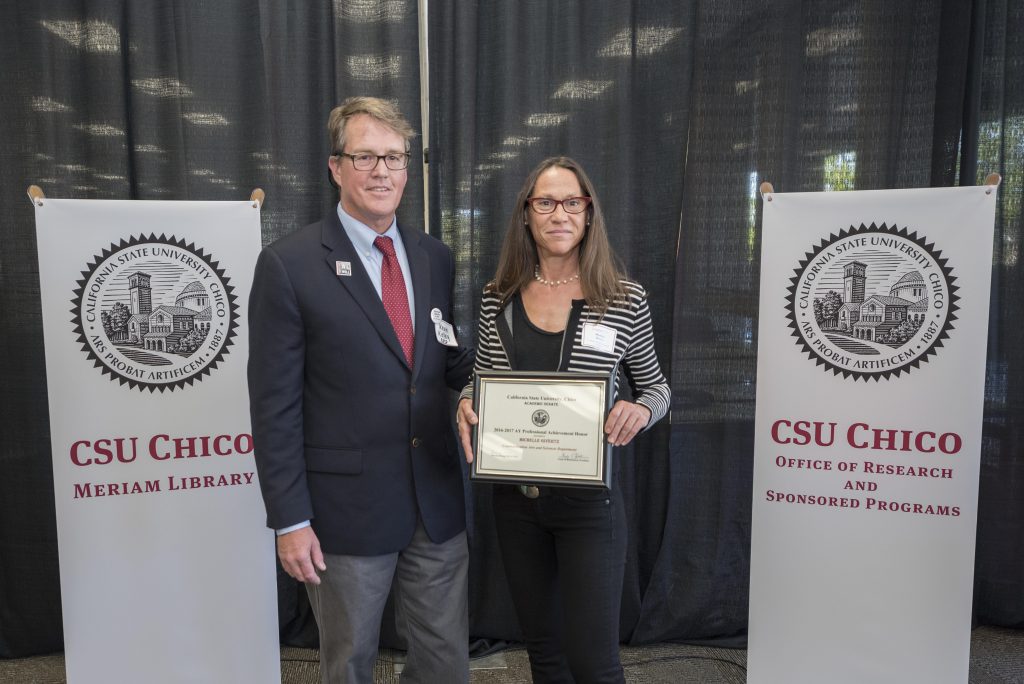 Michelle Givertz, a professor in the Department of Communications Arts & Sciences, holding her Professional Achievement Honors award and stands next to Kevin Kelley, interim associate vice president of the Office of Research and Sponsored Programs.