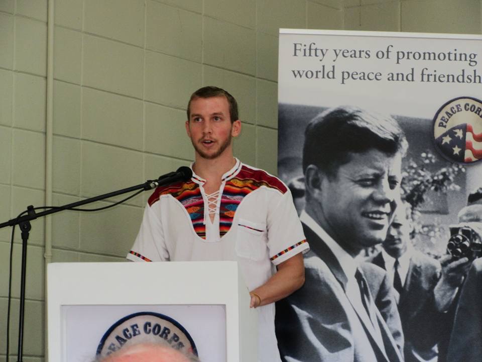 Trevor Johnson speaks at a podium next to a Peace Corps poster of John F. Kennedy.