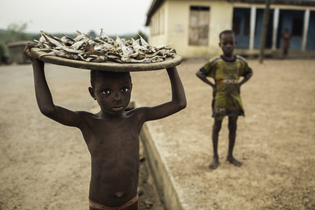 A shirtless Ghanan child stands with a platter of fish resting on his head.