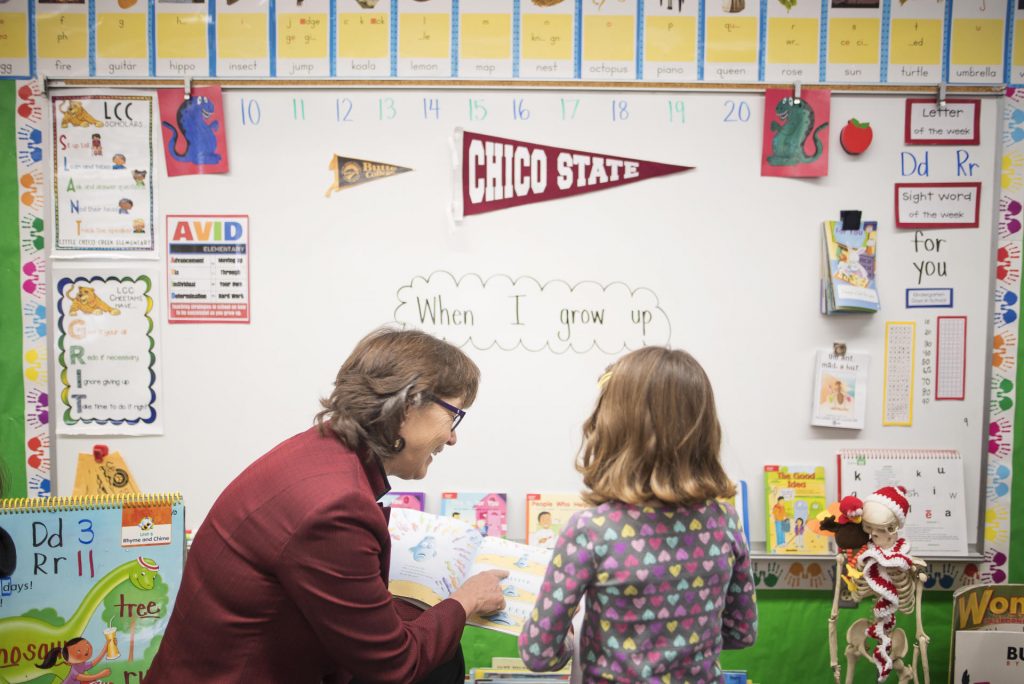 Hutchinson looks at a book with a young student. In the background a Chico State pennant hangs on the whiteboard