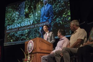 Dylan Gray speaks at a lectern next to other seated speakers on stage in Laxson Auditorium during the President's Fall Convocation.