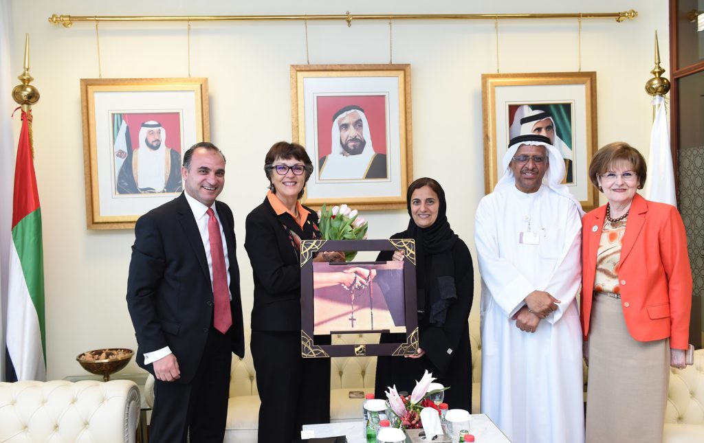President Hutchinson and Sheikha Lubna stand with other dignitaries holding the framed photo of hands holding rosary beads and beads of Islam.