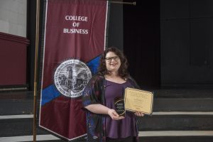 Elizabeth Konecny smiles and holds an award, standing on front of a banner reading College of Business.