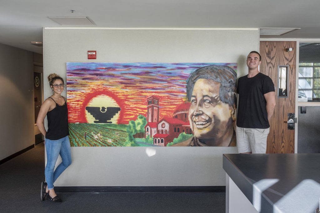 Two students stand on either side of a mural of Cesar Chavez. The mural shows a sunset, a black eagle logo, Chico State buildings and a field.