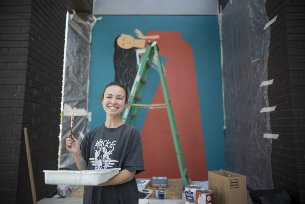 Autumn Robertson smiles, holding a paint tray, in front of a mural of a woman.