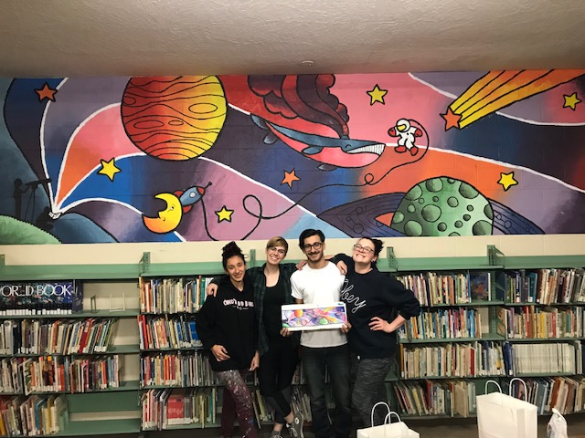 Four students stand in front of a bookshelf and a mural of space-themed art.