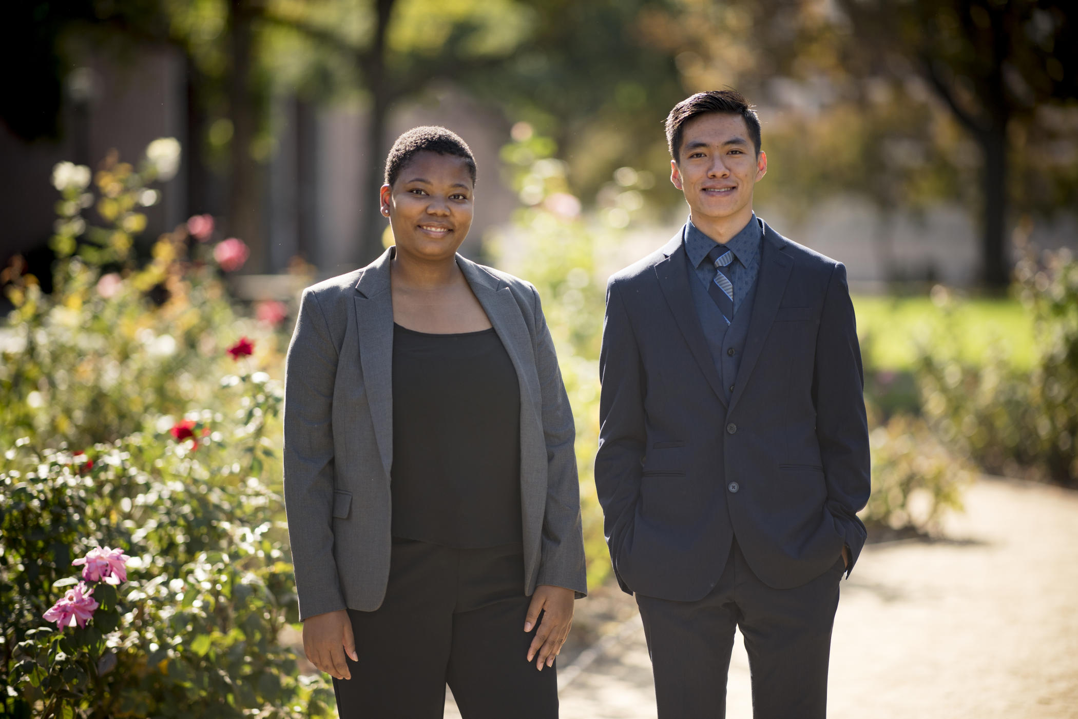 Two students standing in the rose garden, wearing jackets, pants and other professional attire.