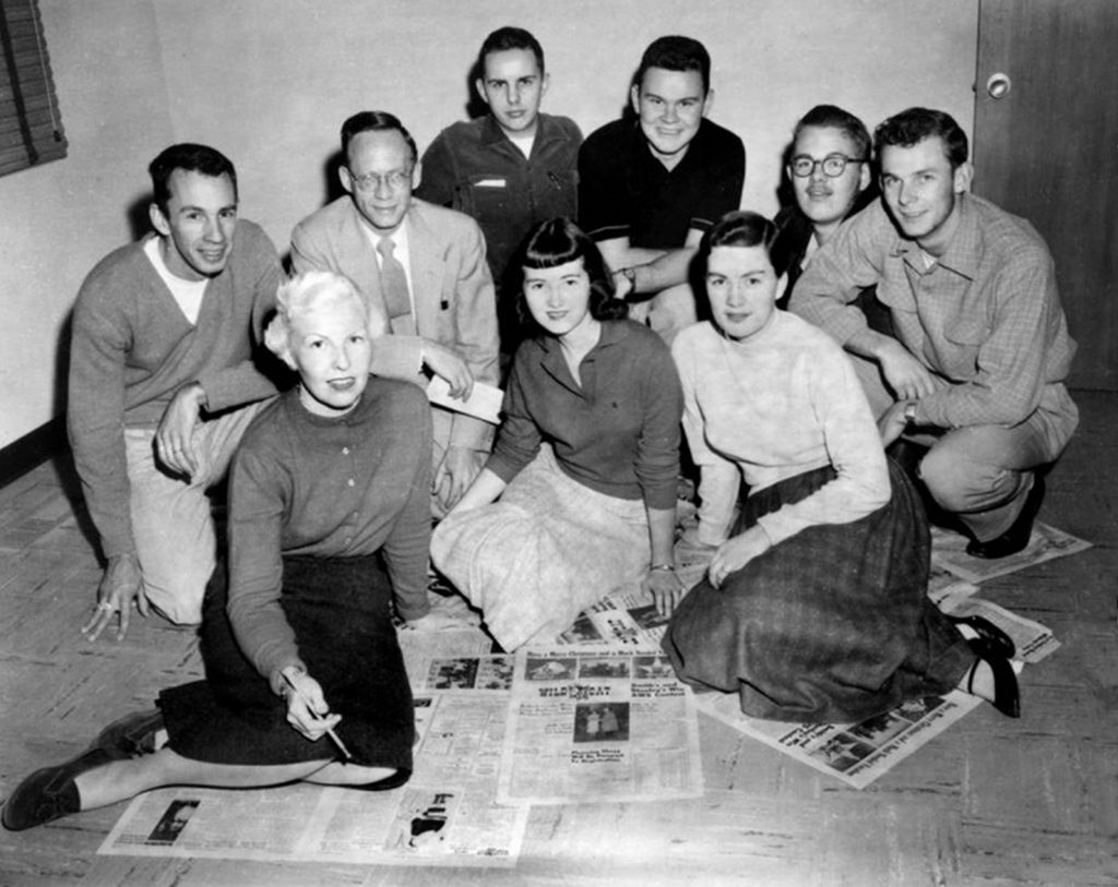 Black and white photo of Faye kneeling on a collection of Wildcat newspapers surrounded by a team of students in collared shirts or long skirts customary to the time.