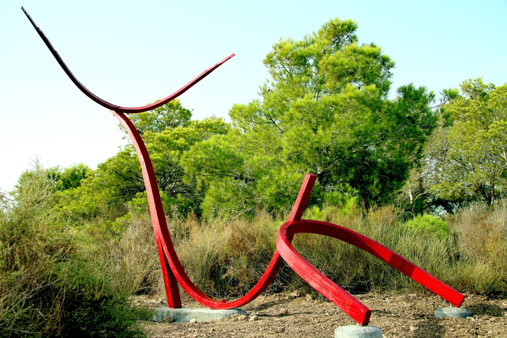 A red sculpture located in the outdoors with trees surrounding it is three U-shaped metal pieces connected 