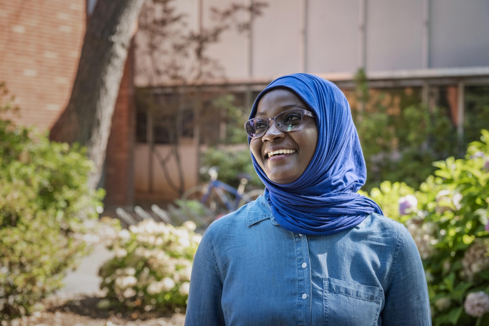 A teacher from Ghana smiles in front of classrooms, wearing a denim top and bright blue scarf on her head.