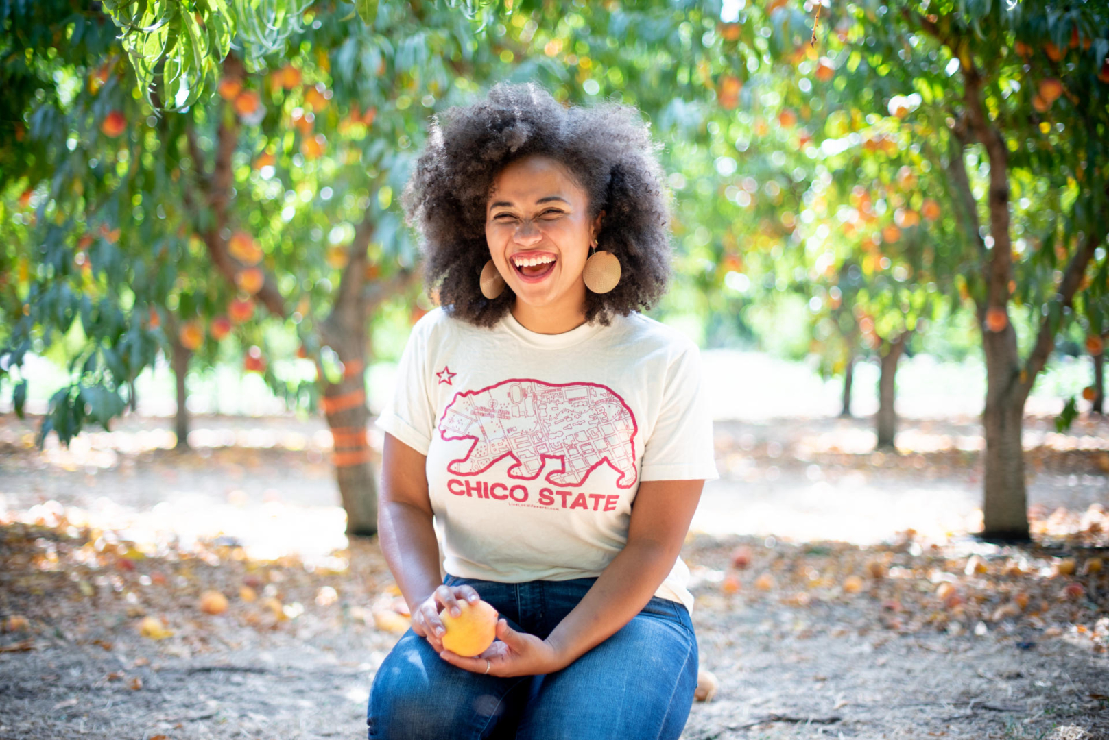 Portrait of Breanna Holbert laughing while sitting amidst peach trees.