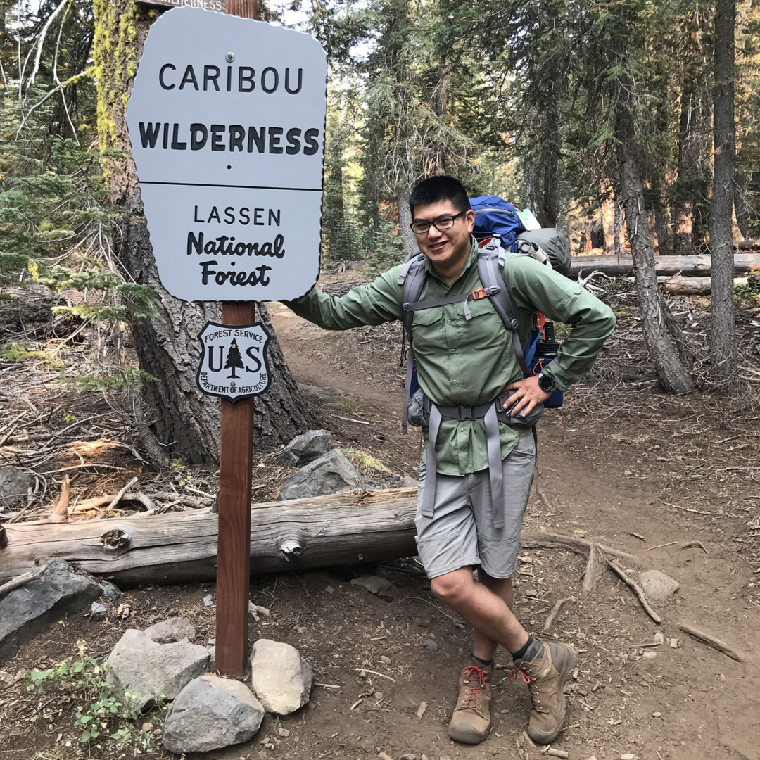 Chang leans against a sign and wears backpacking gear and his backback.