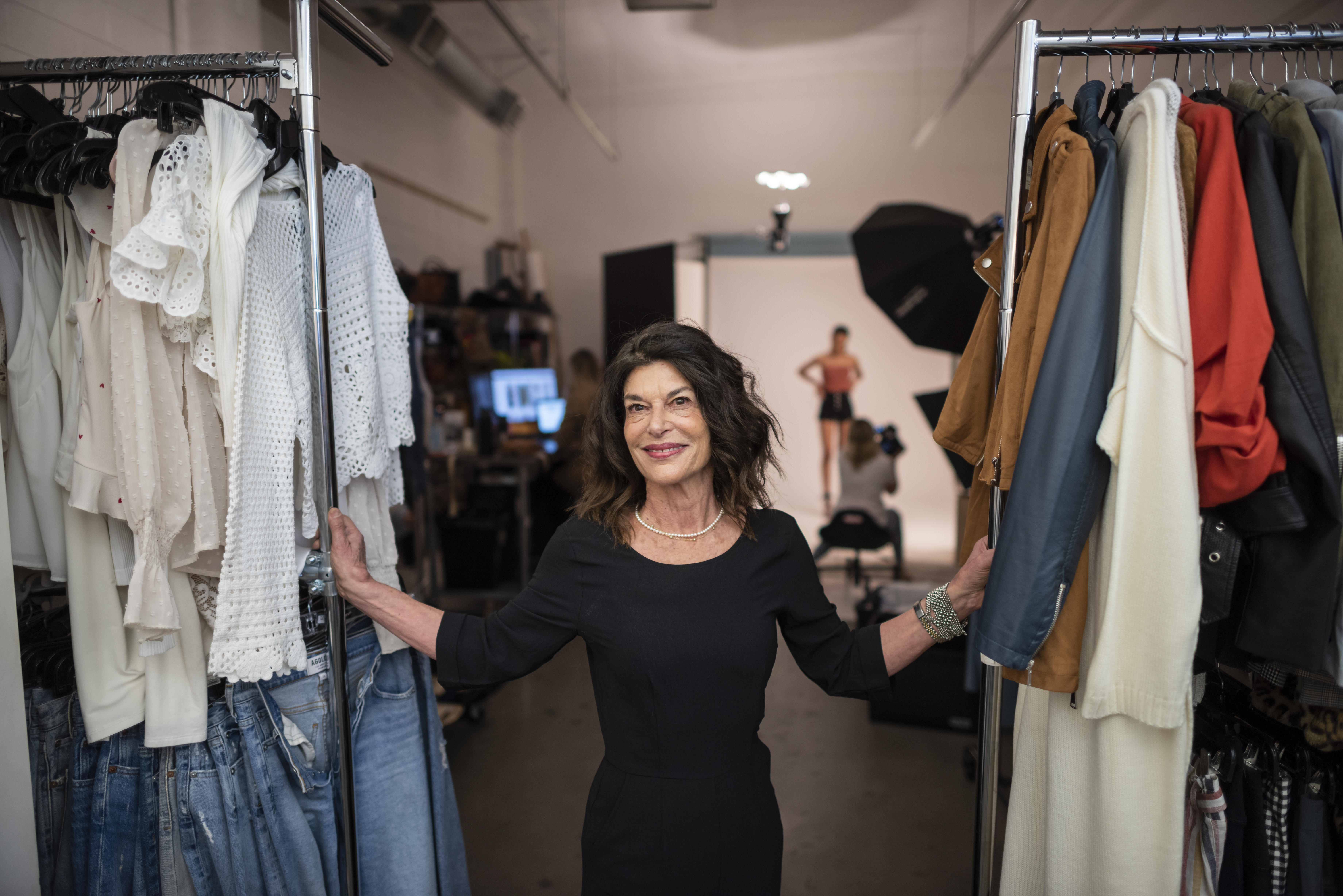 Debra Cannon stands between clothing racks while a fashion photoshoot takes place behind her.