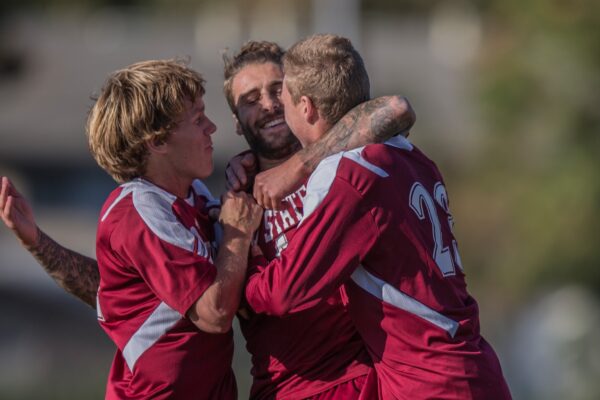 Three men's soccer players embrace in celebration after a game-winning goal.
