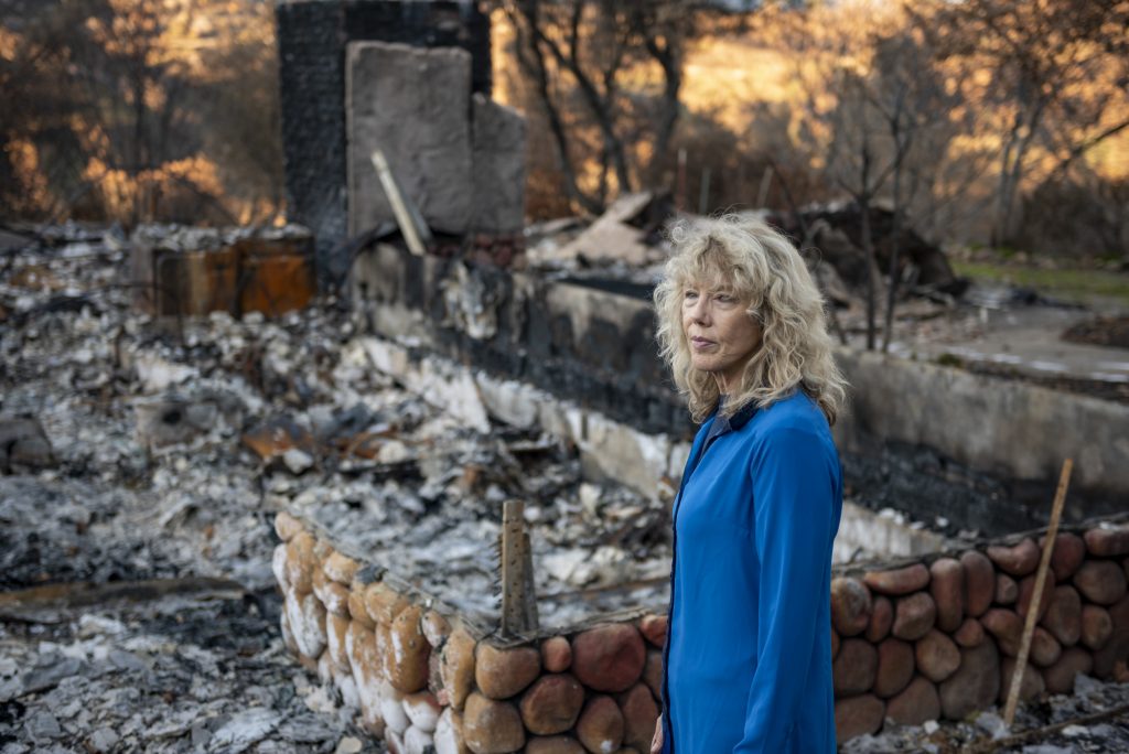 Cindy Wolff stands in front of the ashen rubble of her home, with a rock foundation all that is left.