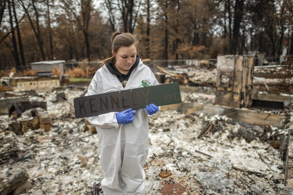 Wearing a protective suit for searching the remains of her home, Kelsea Kennedy holds a charred "Kennedy Avenue" street sign that she found in the rubble.