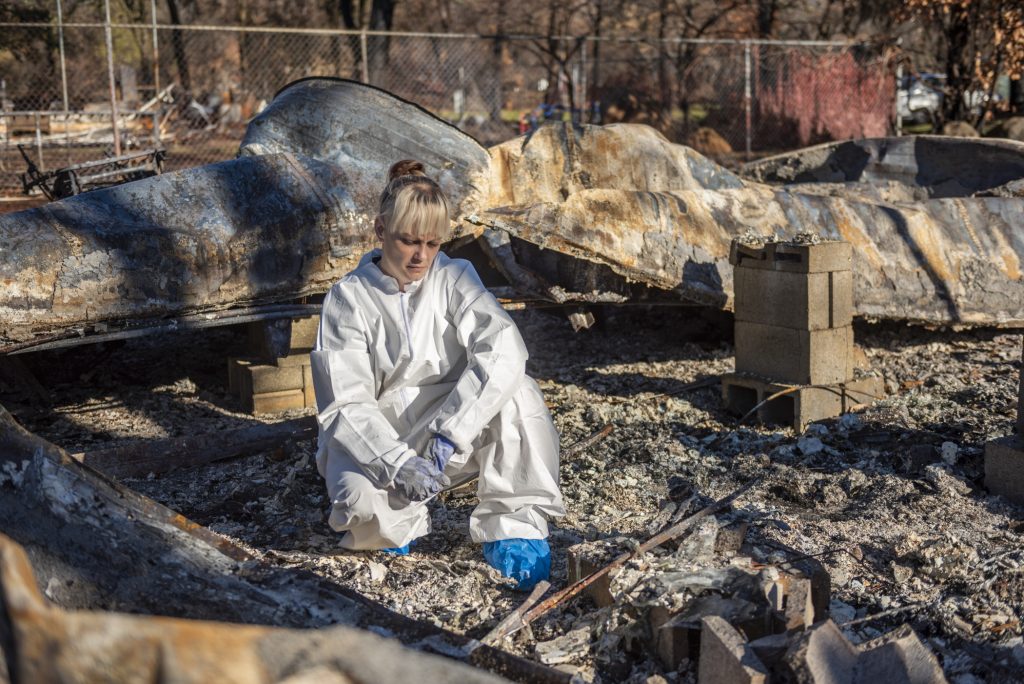 Wearing a protective suit for searching through rubble, Sabrina Hanes crouches in the ashes of the mobile home she was renting and braces herself not to cry.