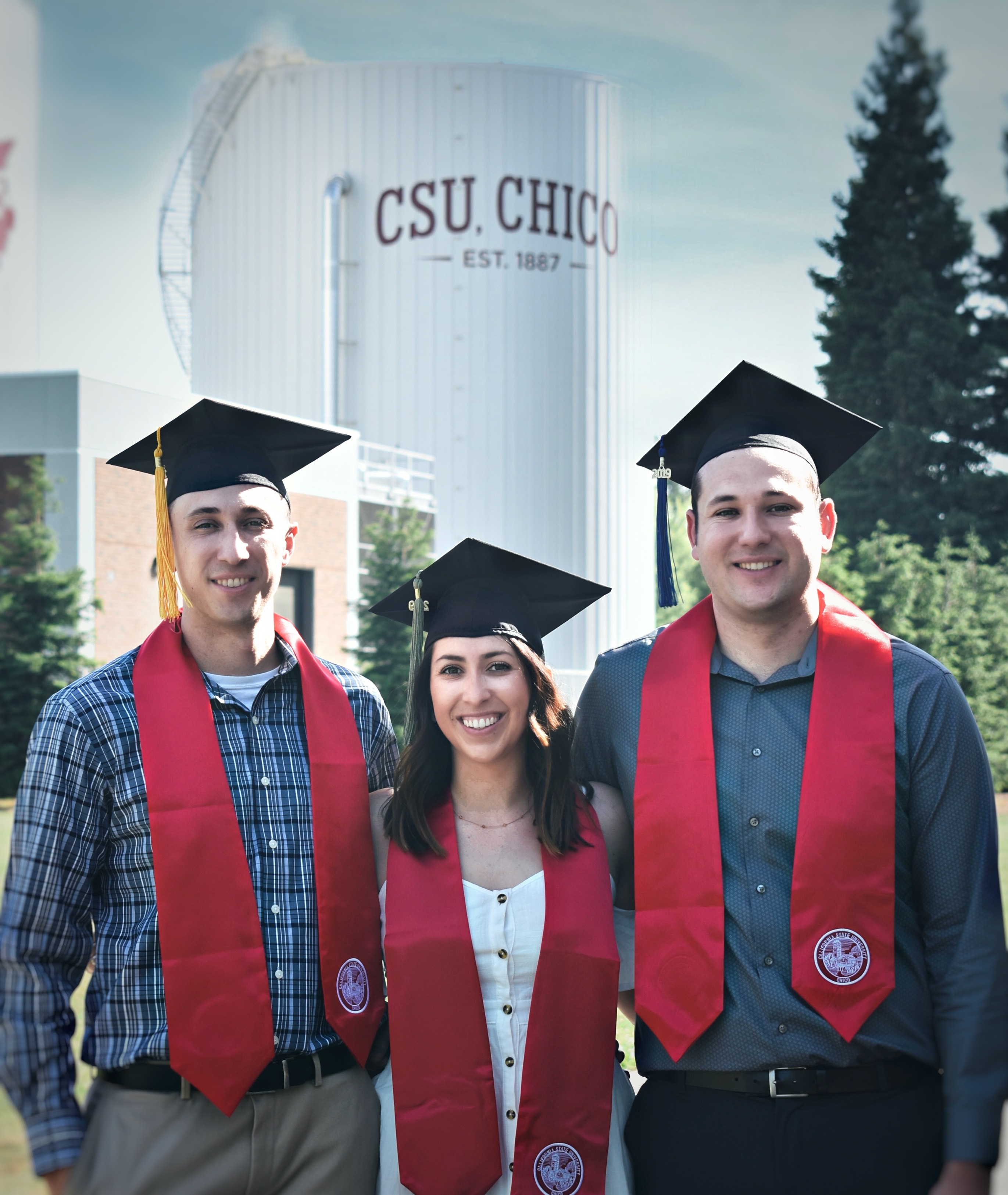 Siblings Nick, Kayla, and AJ Gonzales wear their Chico State stoles as they stand in front of the boiler-chiller towers that say "Chico State."