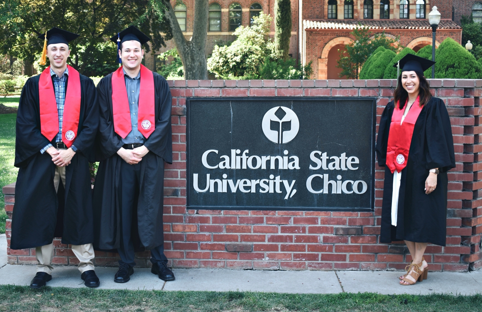 AJ, Nick, and Kayla Gonzales post in their gowns and wearing their Chico State stoles in front of the California State University, Chico monument sign on the Kendall Lawn.
