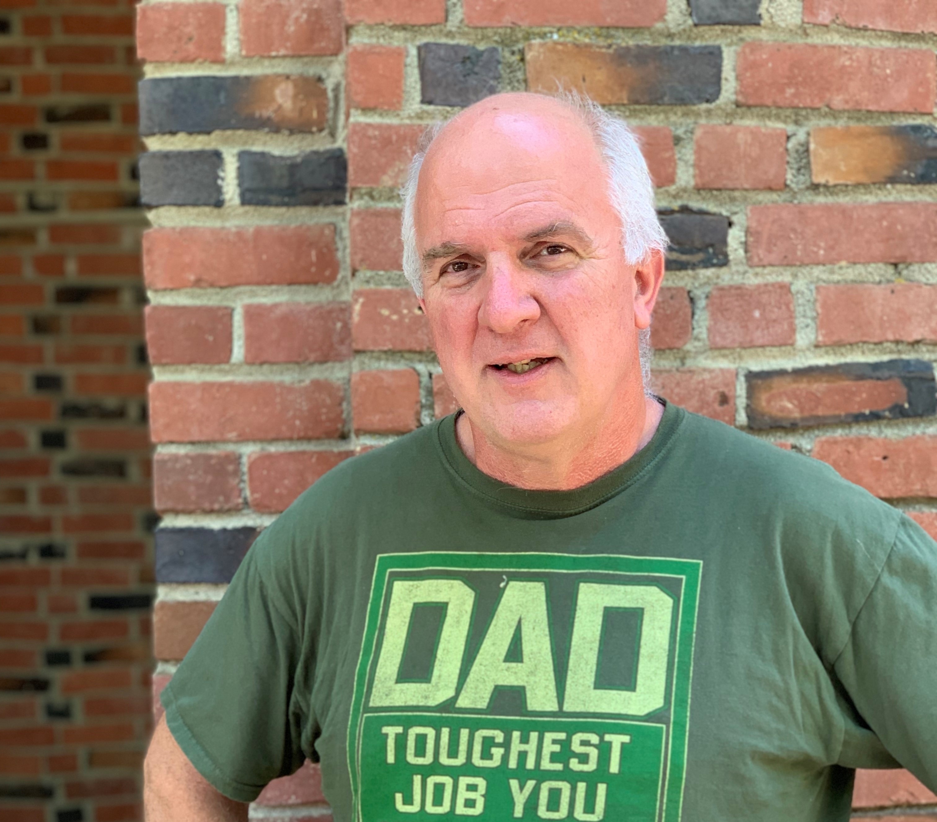 LeRoy Alaways smiles for a photo in front of Kendall Hall wearing a shirt that says "Dad toughest job you will ever love."