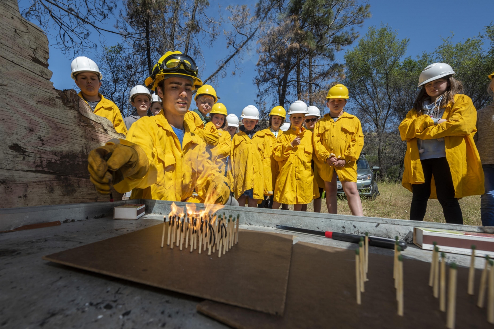 A firefighter lights a grove of matches standing upright on a flat surface as fourth graders watch behind him in protective yellow clothes.