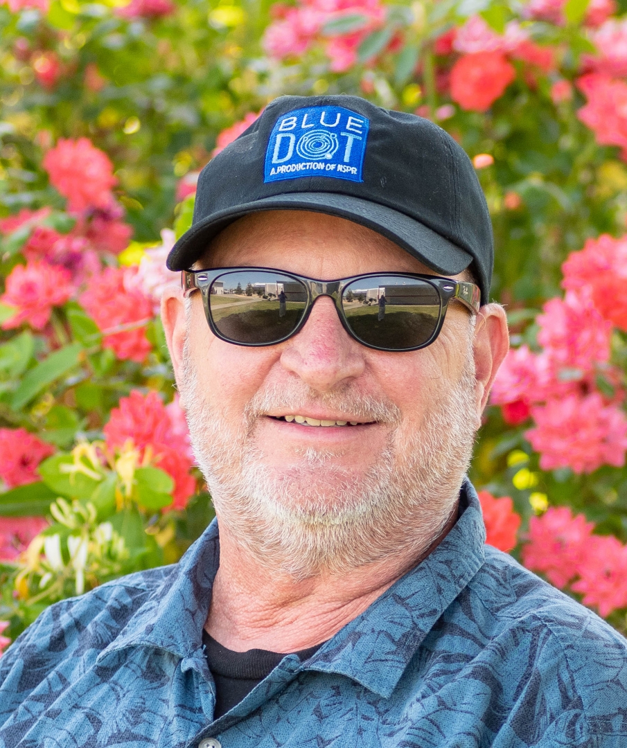 Dave Schlom wears a baseball cap and poses in front of colorful flowers.