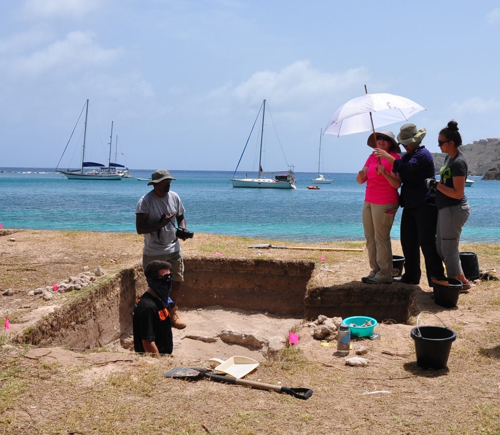 A group of researchers stand in an excavation site with the ocean in the background.