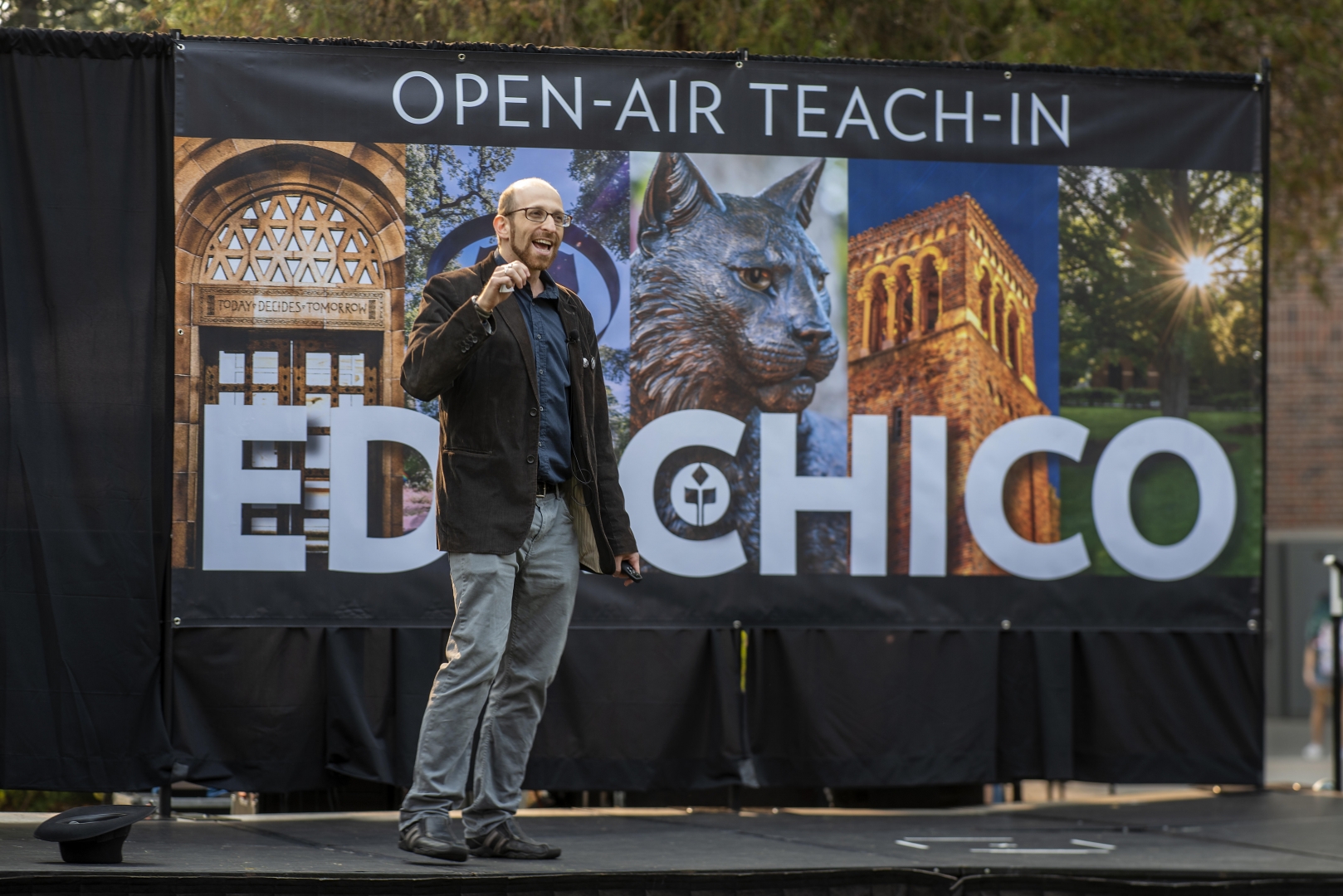 Asa Mittman stands on stage in front of an EDXCHICO poster, speaking to a crowd.