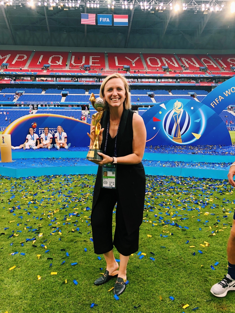 Molly Downtain smiles and holds the World Cup trophy on a soccer field littered with confetti after the World Cup.
