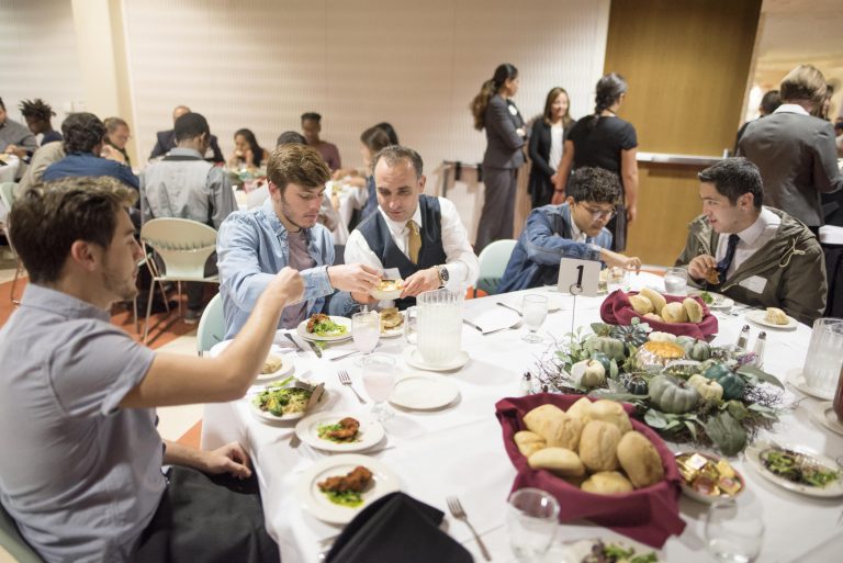 Students and faculty sit around a dining table enjoying a meal.