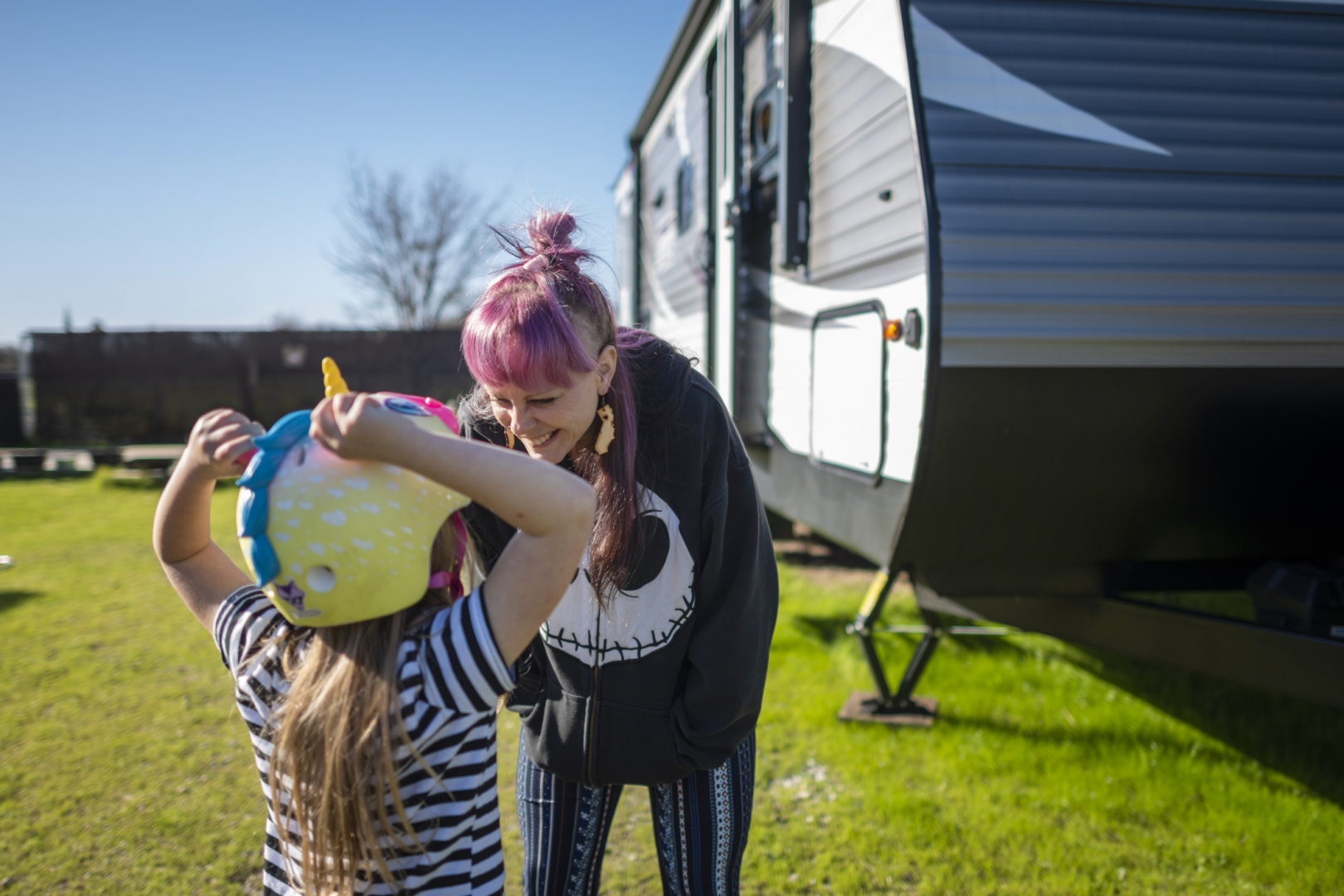 Sabrina Hanes stands outside her trailer and leans over to smile at her daughter as she adjusts her unicorn bike helmet.