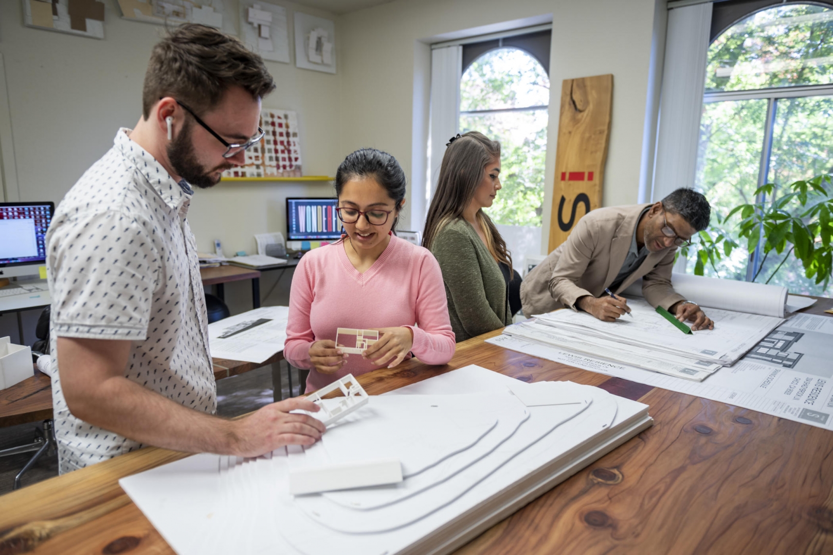Two students stand at a long table to discuss 3-D mockups of houses while another student and Rouben Mohiuddin review paper designs nearby.