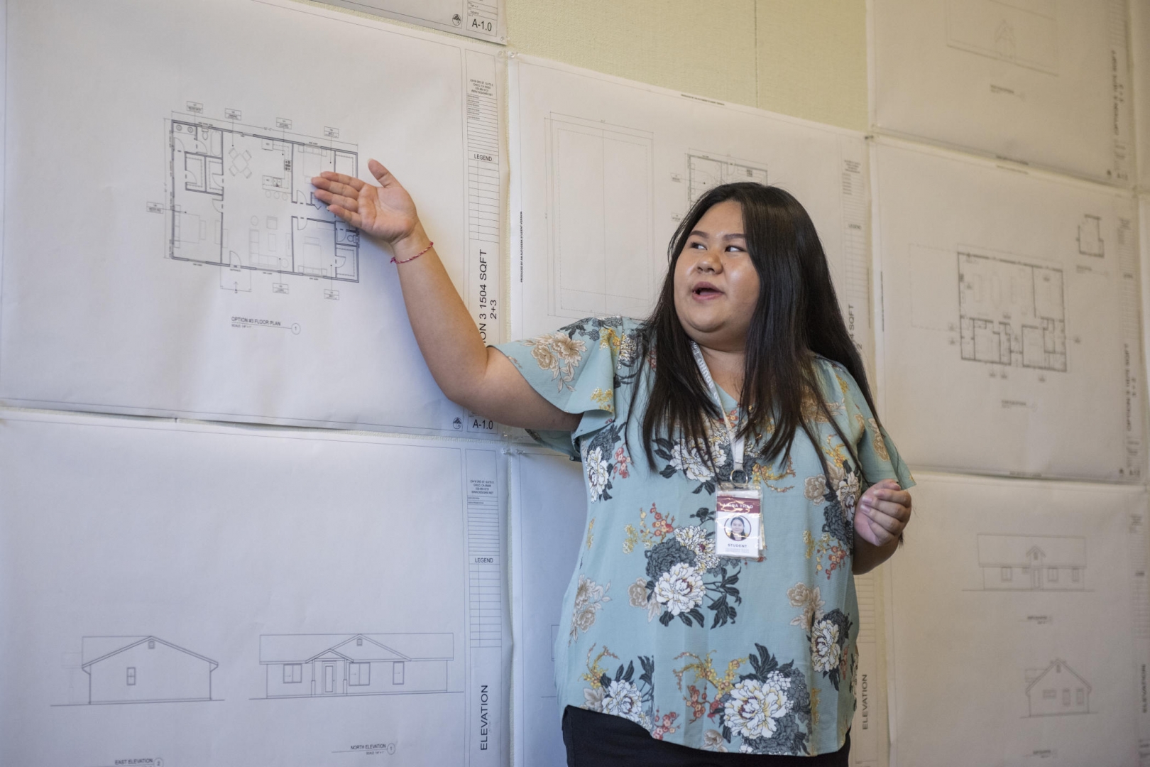 Linda Thao gestures to designs of houses sketched onto paper that is on a wall as she talks about them.
