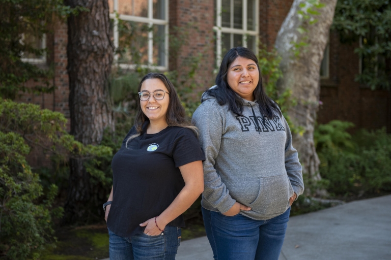 Two female college students stand back to back in front of trees.