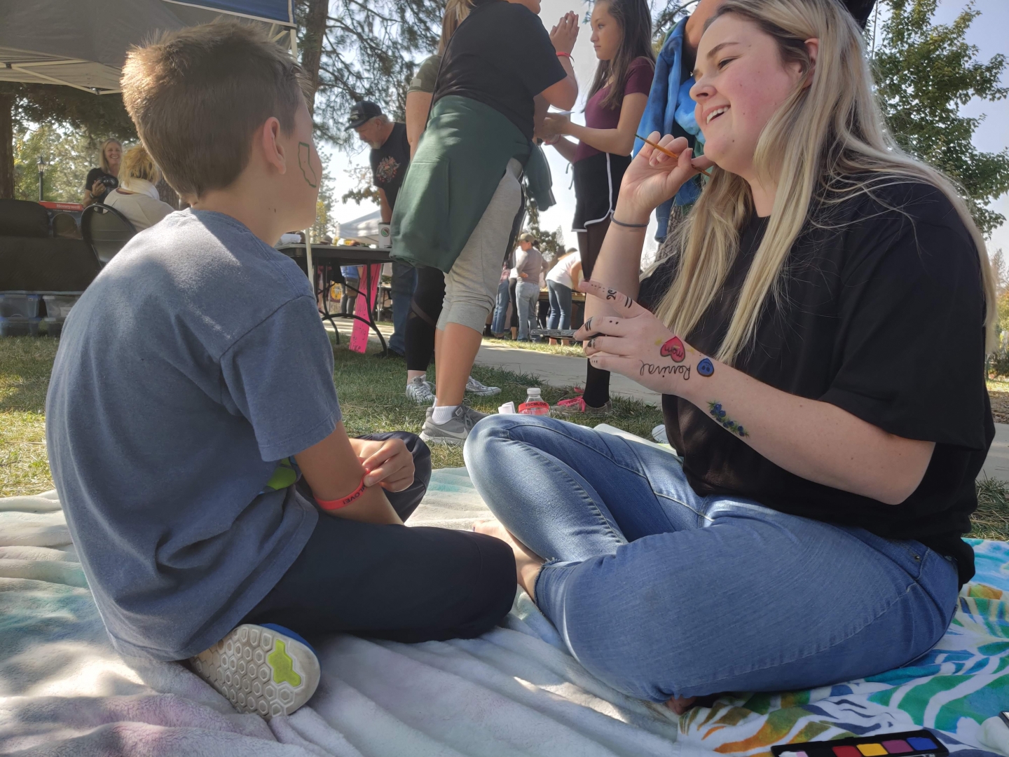 A boy sits to have the outline of California painted on his cheek by a smiling young woman while they sit on blankets on the grass.