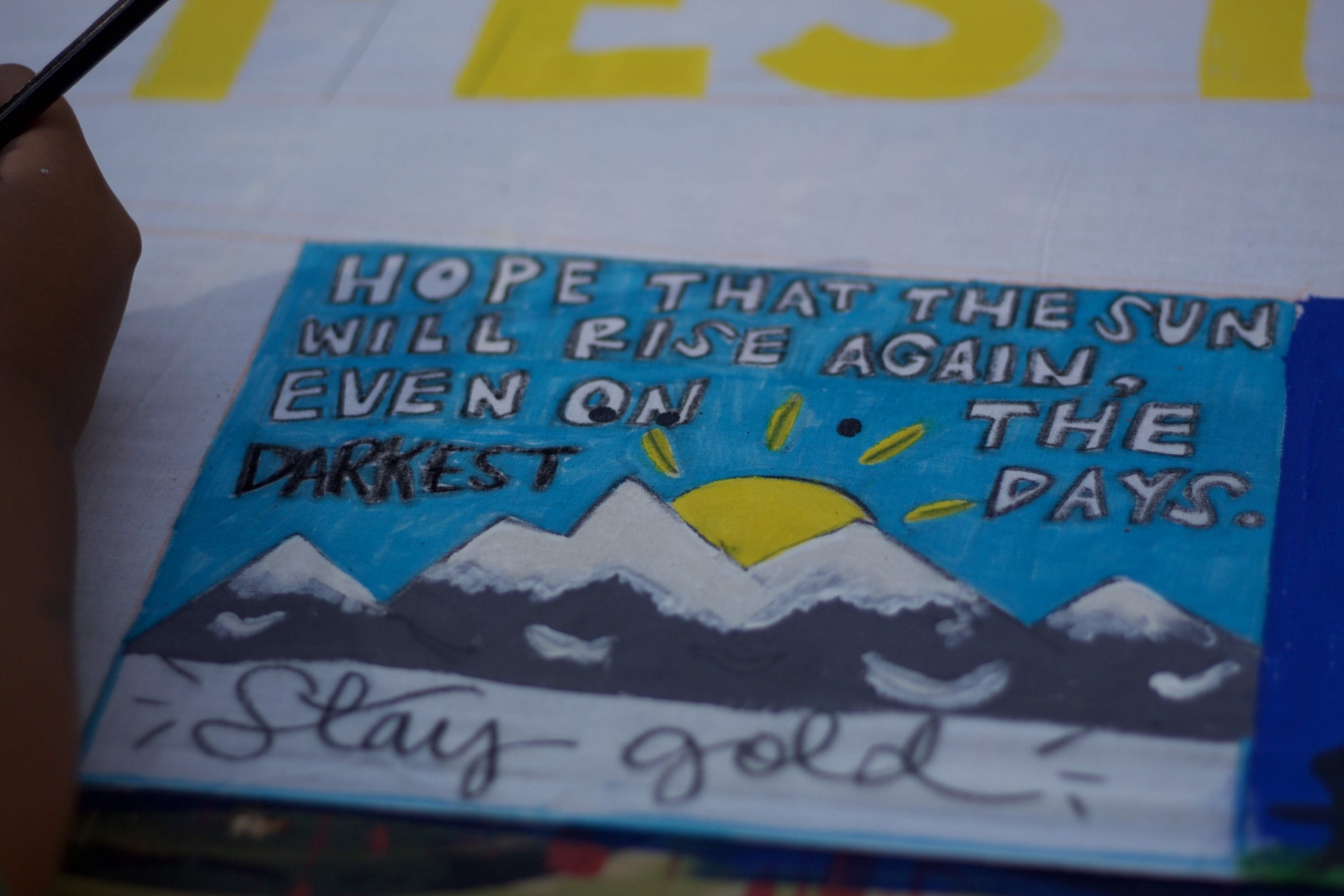 The phrase "Hope that the sun will rise again, even on the darkest days. Stay gold" is painted on a backdrop of snow-capped mountains with a sun rising behind them.