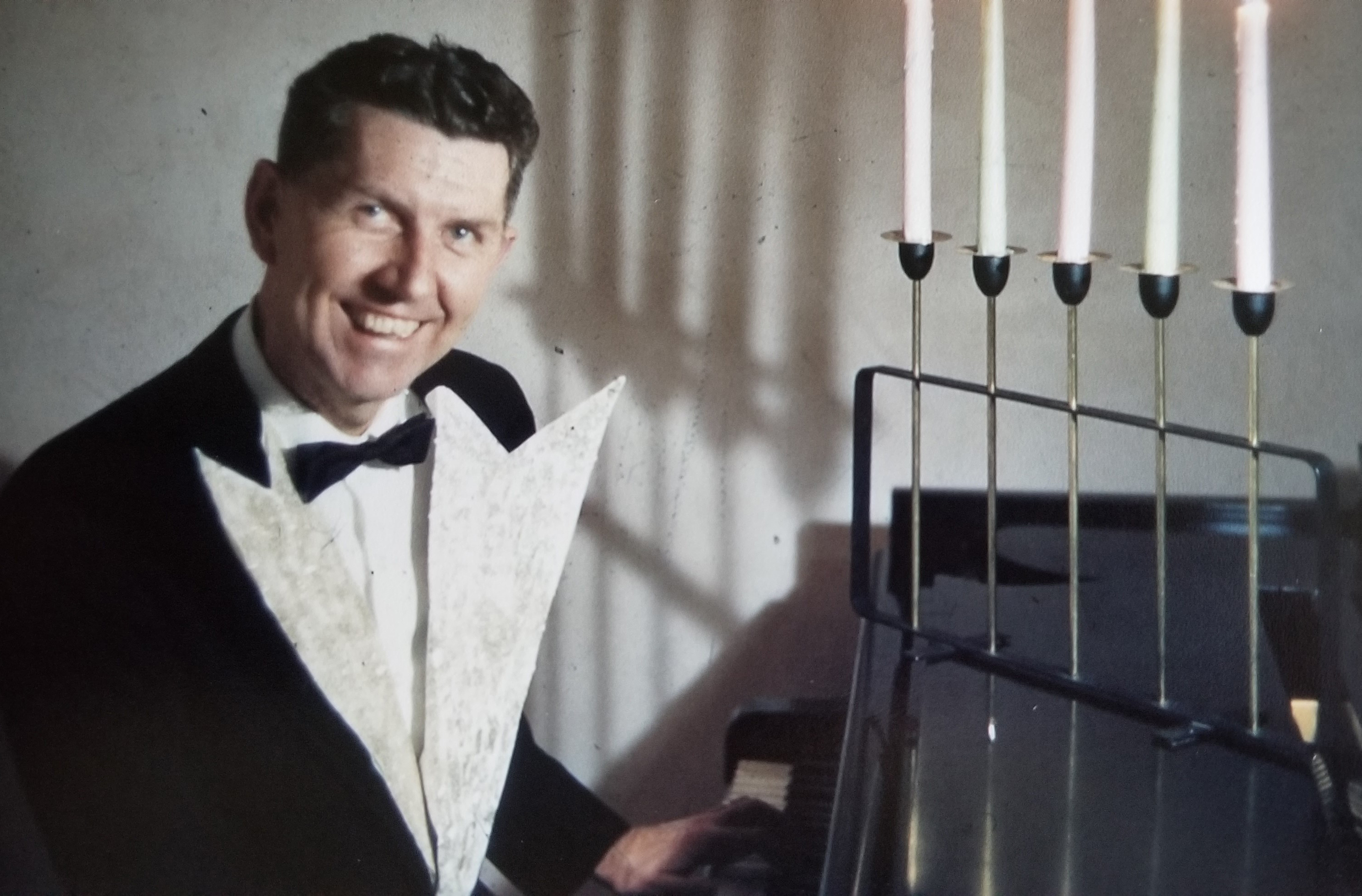 Robert Laxson sits at a piano while wearing a tuxedo.