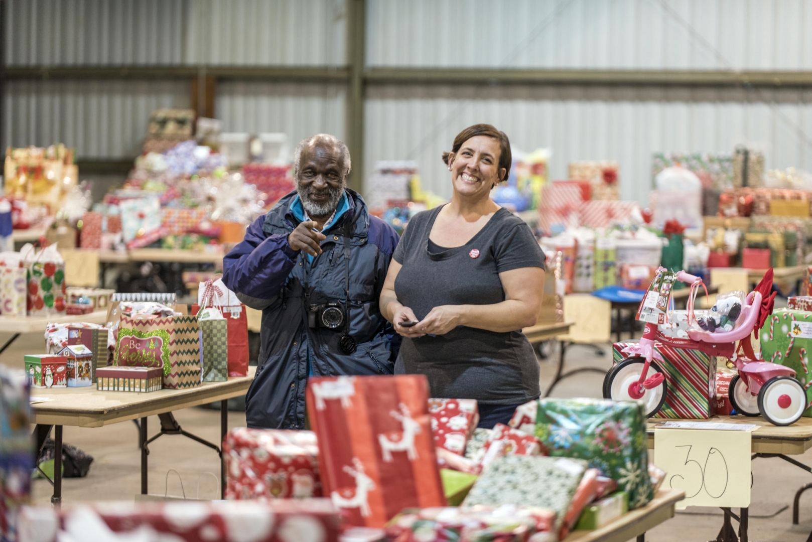 Two staff members smile while standing amid tables filled with wrapped gifts.