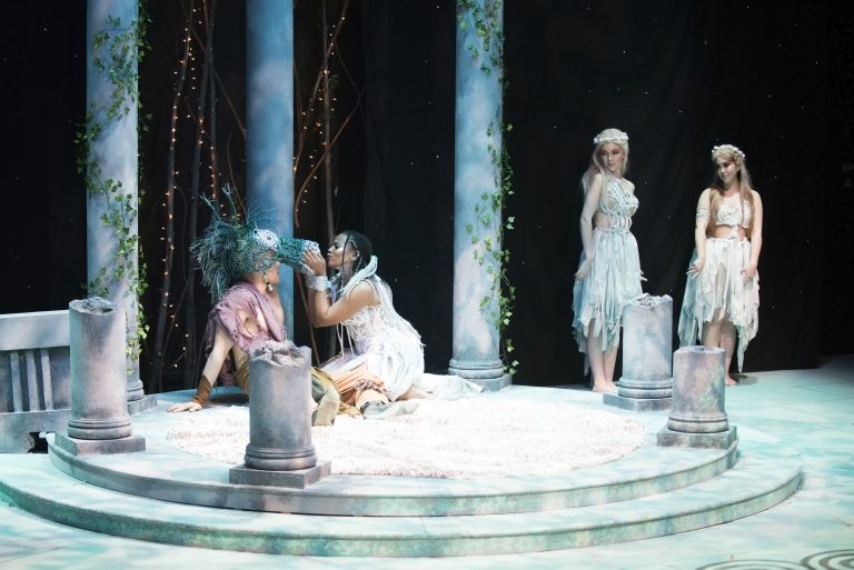 Actresses in long white dresses rehearse on an elaborate set design.