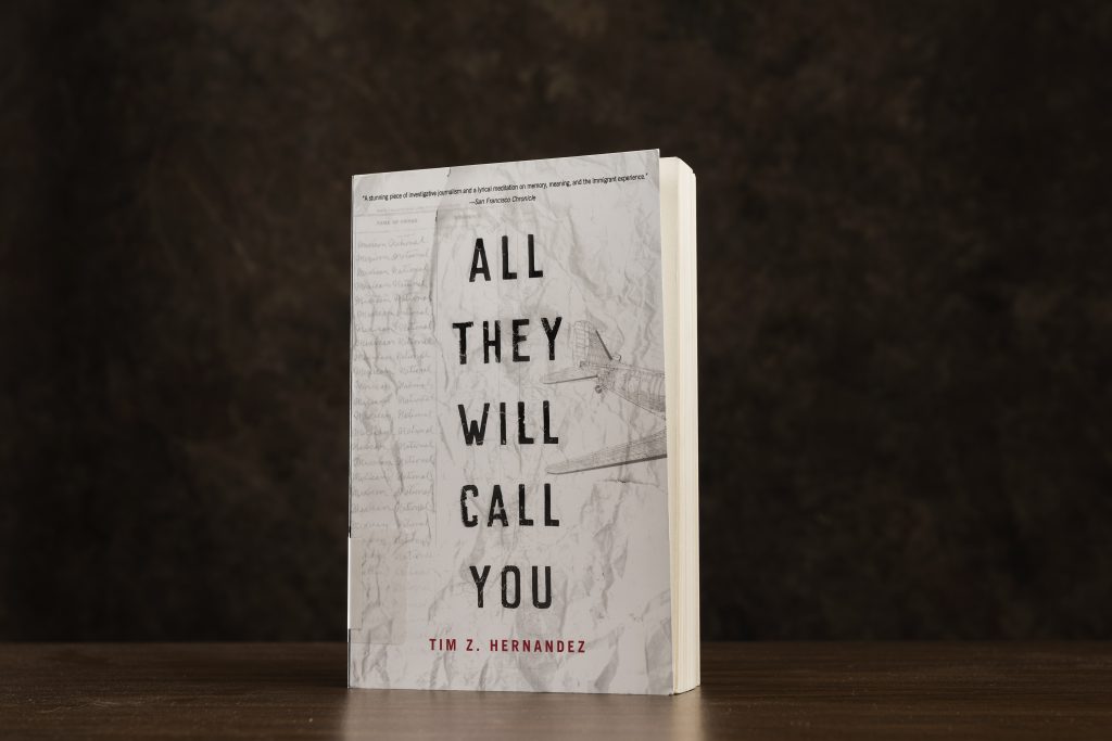 The book "All They Will Call You," with a white cover, is silhouetted against a black background