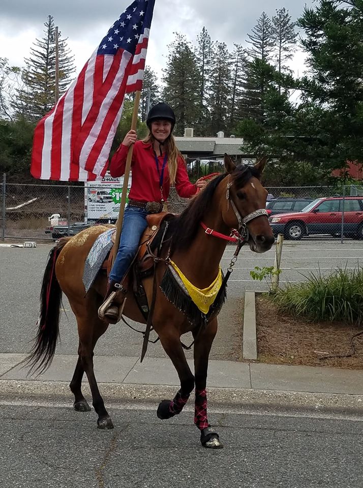 Cassidy Sabral holds a large American flag while sitting on the back of a horse.