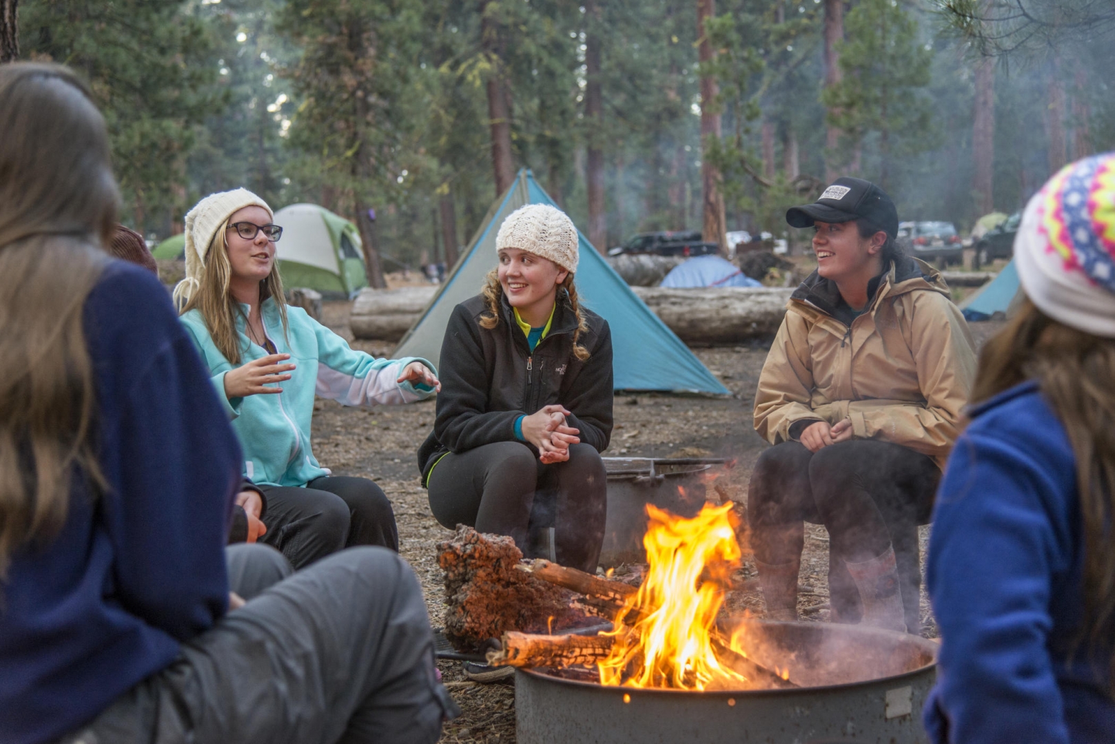 Students tell stories and laugh around a camp fire, with their tents behind them.