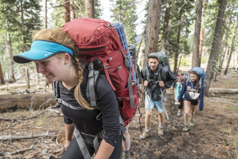 A student hikes hunched over with a heavy backpacking pack on her back as other students follow behind in a forest.