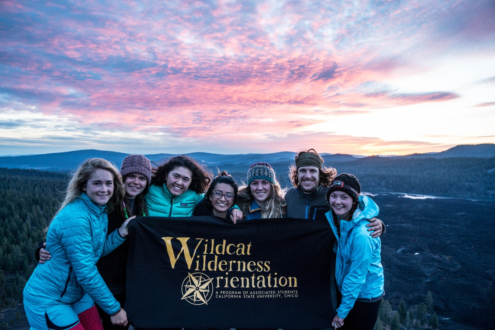 Students hold a Wildcat Wilderness Orientation sign at the top of a mountain with a colorful sky behind them.