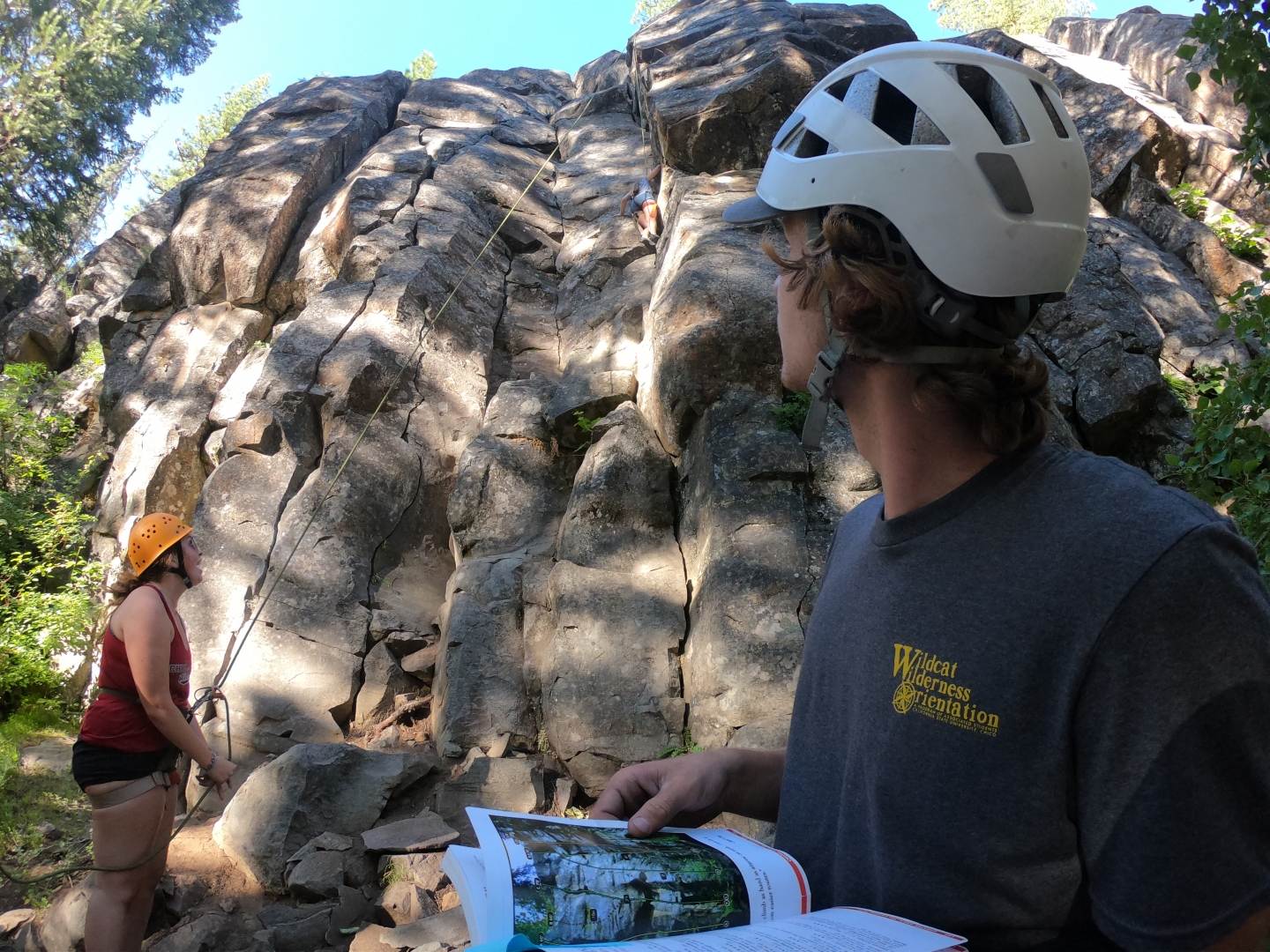 A student reads a guide book on rock climbing as students wearing helmets ascend a rock wall behind him.
