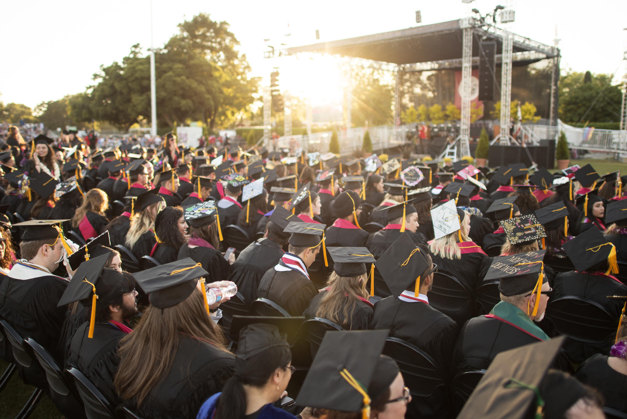 Graduates in cap and gown sit in anticipation of Commencement as the sun sets behind the stage