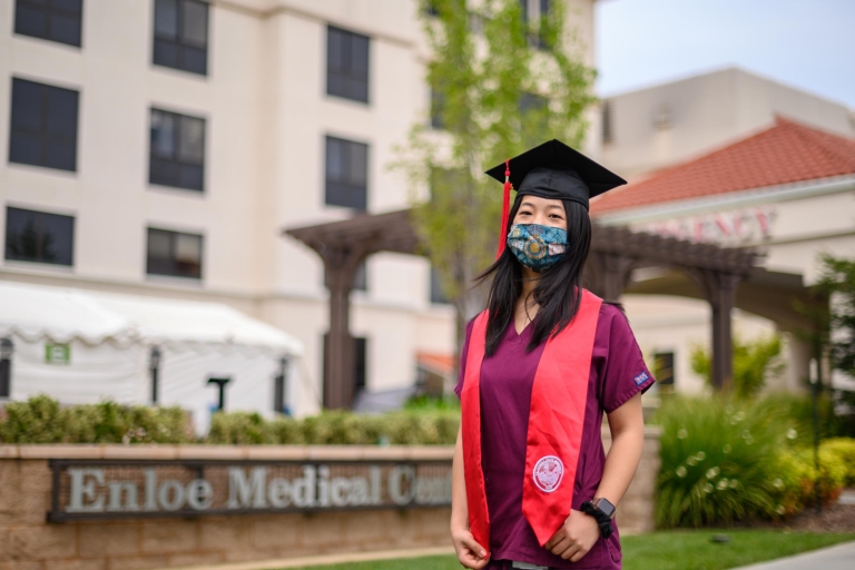 Amy Peet stands in front of Enloe Hospital wearing a face mask and her commencement attire.
