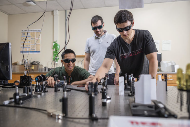 Three men in protective eyewear do laser testing over a table.