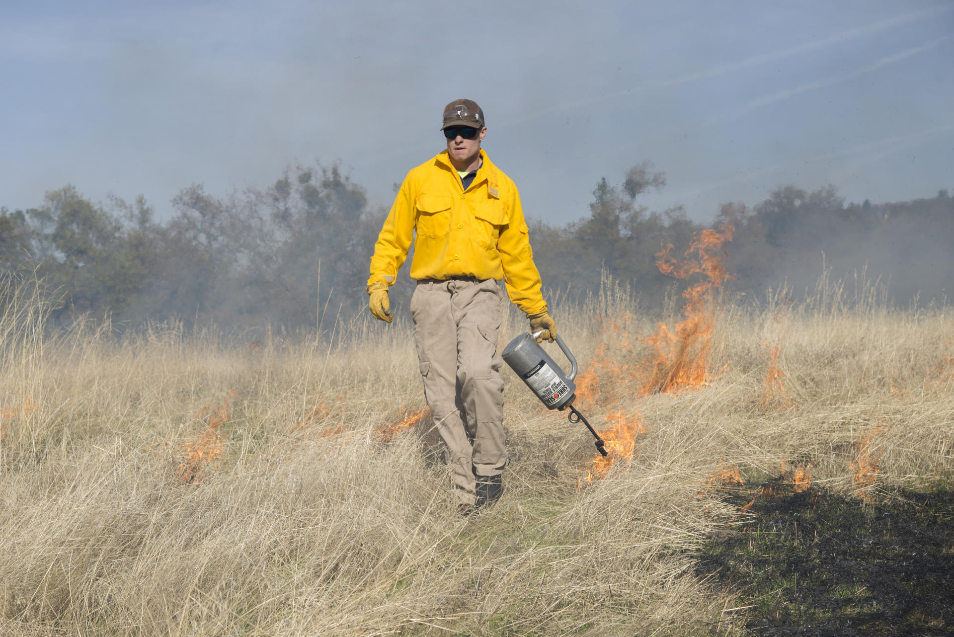 A student walks with a drip torch lighting off fire in a dry field.